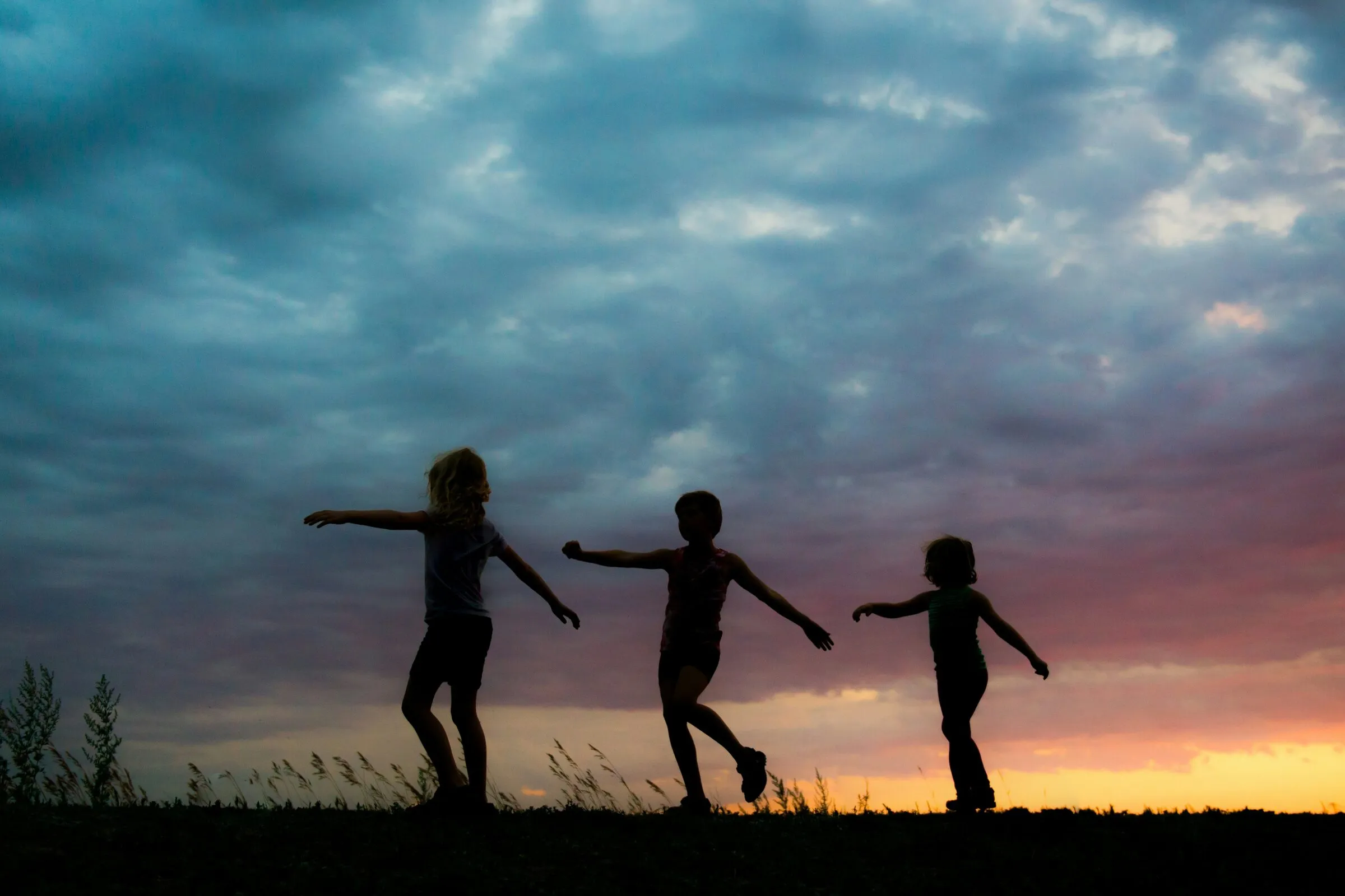 Three children dancing on a hill against a stormy sunset sky