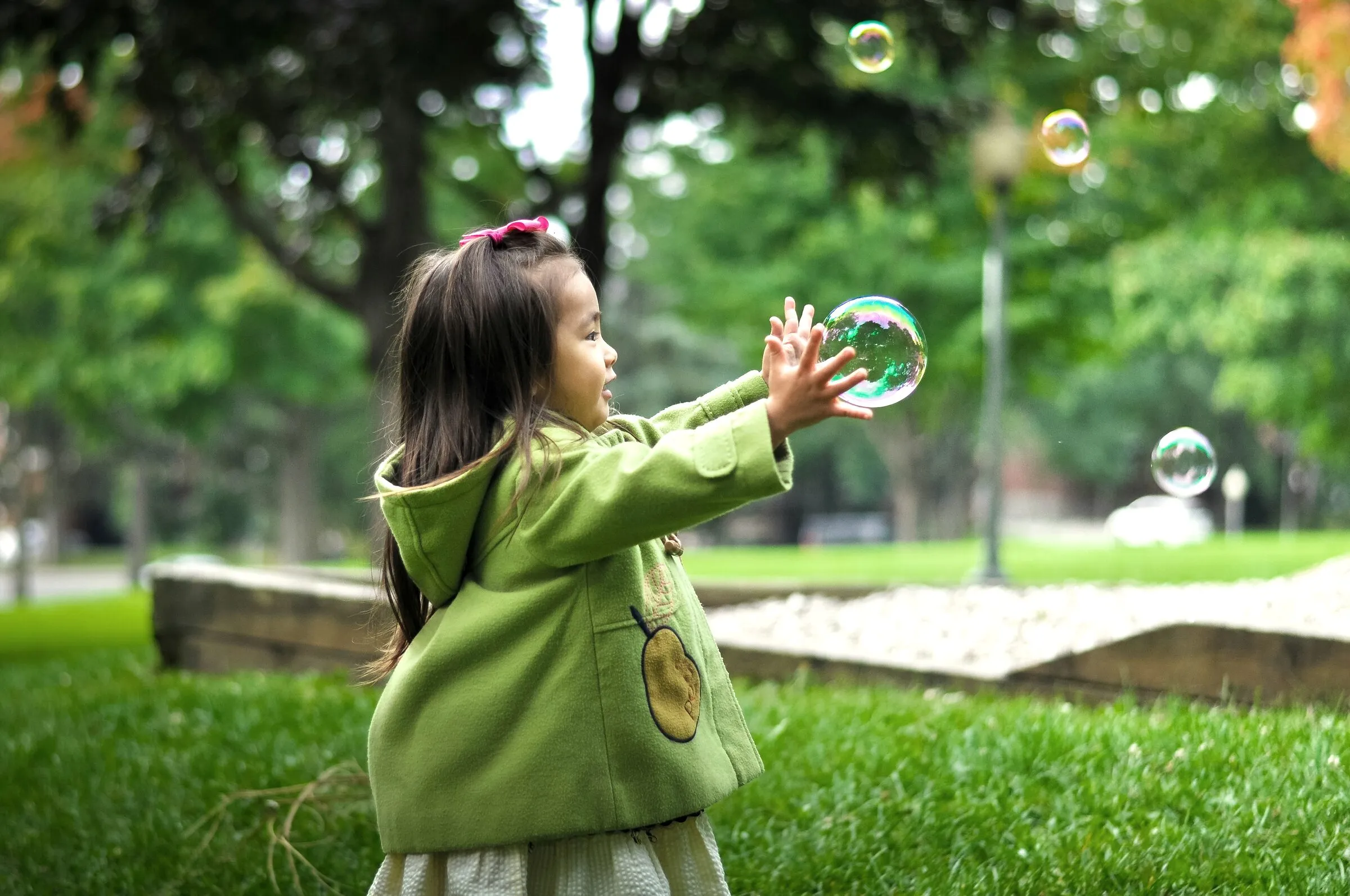 A little girl in a green coat reaching out to catch a soap bubble in a park