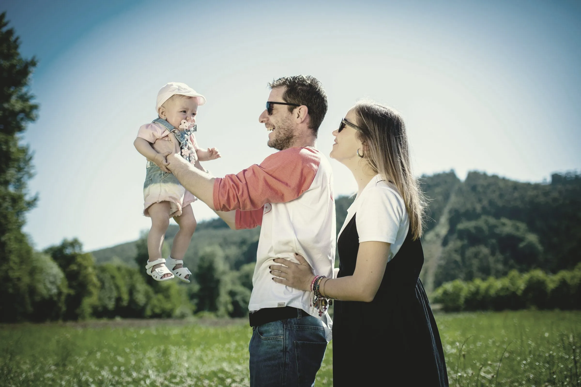 A smiling father lifts his baby daughter into the air as her mother looks on, in a sunny meadow